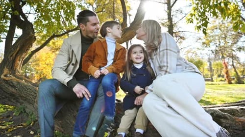 Happy family in an autumn park. Mother, father, son and daughter sitting on a tree trunk, mom kissin