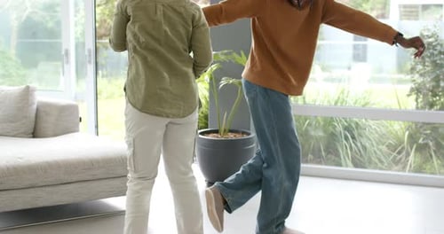 Casal afro-americano praticando passos de dança, braços estendidos por portas de vidro deslizantes na sala de estar