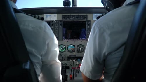 Two pilots inside the cabin of a small plane, filmed from behind while preparing to land.