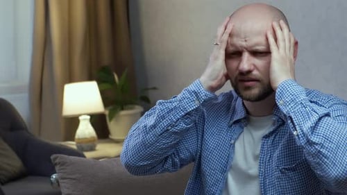 Ein glatzköpfiger Mann mit Bart berührt seinen Kopf und blinzelt vor Schmerzen