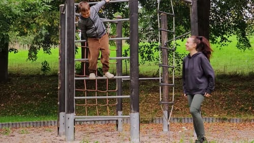 Eine junge Mutter unterstützt ihr Kind beim Aufstieg in ein Fitnessstudio im Dschungel auf einem Spielplatz.