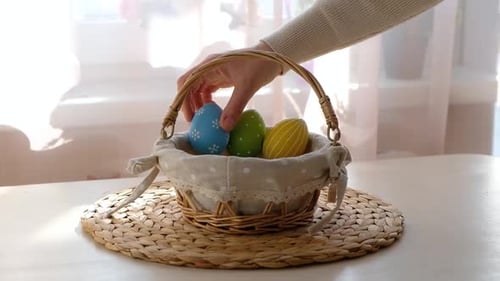 Weibliche Hände legen bunte Ostereier in einen Korb auf einen weißen Holztisch