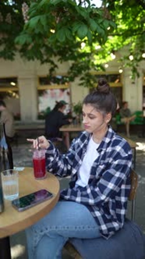 On the Café Terrace a Stunning Young Woman with a Smartphone Chats Away While Enjoying a Cocktail