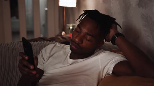 A tired young black male scrolls through his phone while sat on the sofa