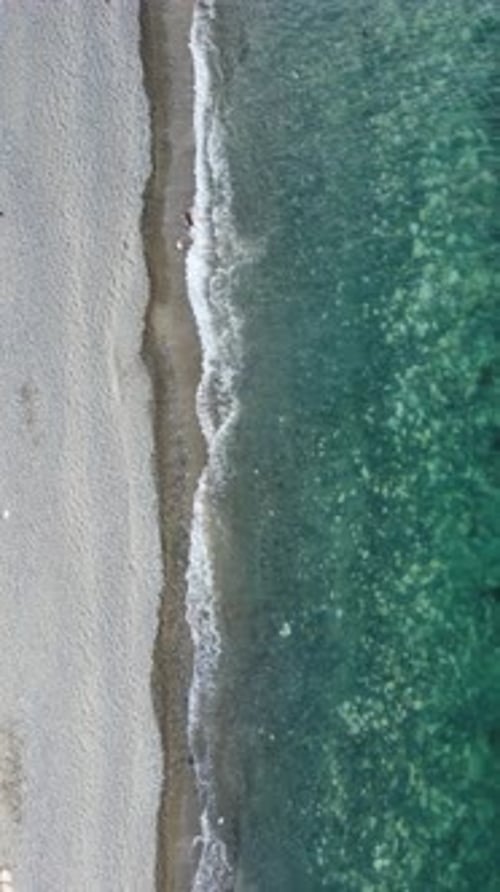 Vertikale Luftaufnahme des Strandes mit Wellen, die die Küste streicheln, Sonnenblendung, kleine Wellen auf der Wasseroberfläche