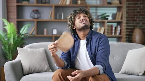 Young adult man is hot while sitting on the sofa in the living room at home. Upset disappointed male