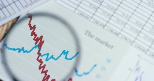 A hand with a magnifying glass passes over a financial newspaper and financial bag with a business
