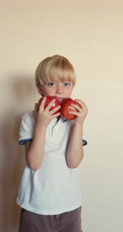 Un niño rubio con una camiseta blanca que sostiene dos manzanas rojas frente a una pared blanca que muestra la hermosa