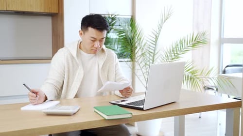 young Asian man counts bills, bank checks, loans or utilities while sitting at home at a kitchen