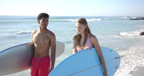 Biracial man and Caucasian woman hold surfboards on a sunny beach