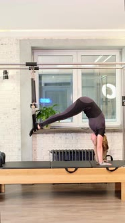 A Woman Performs an Inverted Pilates Pose on a Reformer Tower Holding the Bar with Both Hands The