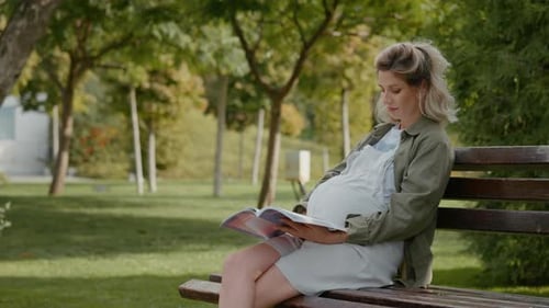 Pregnant female focused on a maternity book while sitting on a bench.