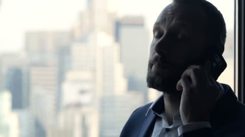 Angry Businessman Talking on Cellphone by Window in Office 30s