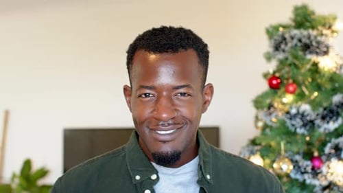 Hombre afroamericano sonriente de pie junto al árbol de Navidad en su casa, sintiéndose alegre