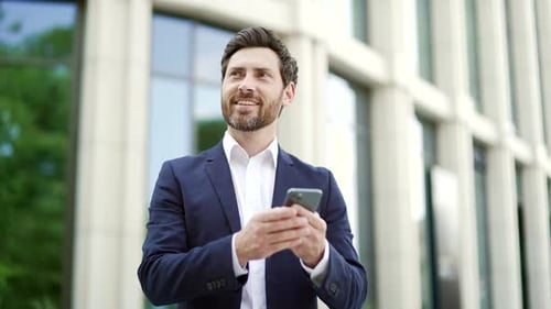 Happy businessman in suit uses mobile phone standing on city street outside near modern office