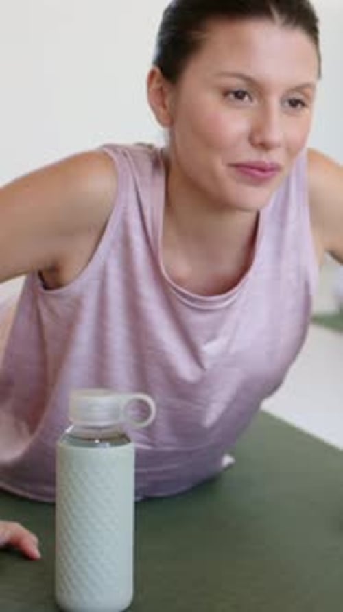Vídeo vertical: Colocando las manos, levantando el pecho, mujer con la blusa rosa haciendo yoga en casa con su pareja, botella