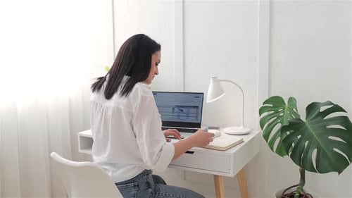 Back View Young Woman Student Sitting at Home at White Desk Using Laptop Writing Notes While