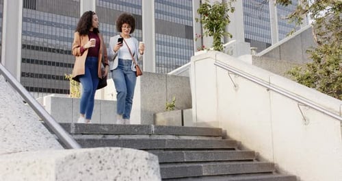 Holding coffee cups, Diverse female friends descending stairs in plaza while swiping smartphone
