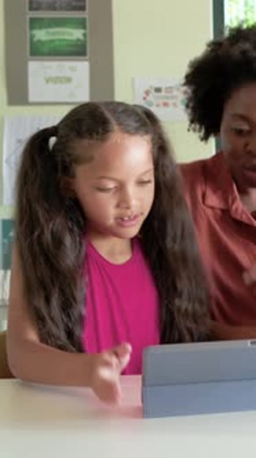 Vertical video: Leaning teacher in rust shirt guiding child at class table, holding blue tablet