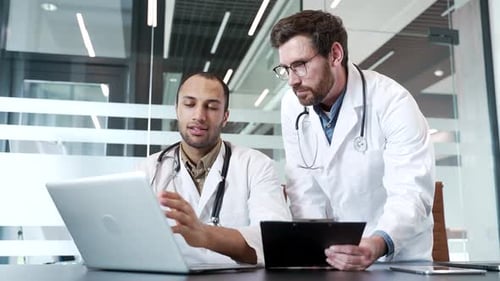 Two doctors in white coats collaborate over laptop and folder while sitting in hospital office.