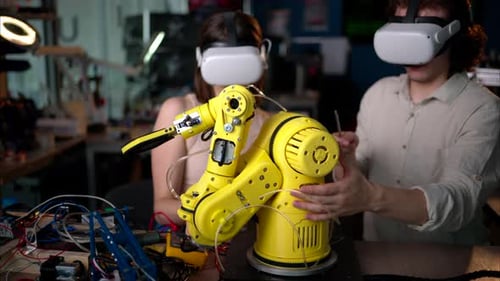 Two young happy engineers with VR virtual reality headsets fixing an yellow robotic arm in the works