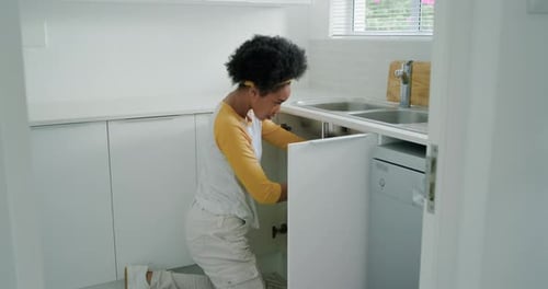 Woman kneeling opening cabinet under sink in kitchen, inspecting plumbing and using toolkit