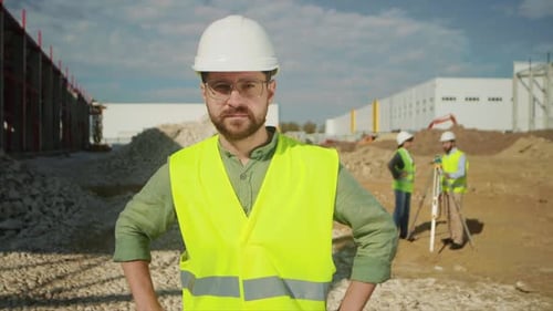 Portrait of Young Bearded Man in Glasses Dressed in Bright Vest and White Hard Hat Looking at Camera