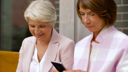 Happy senior women friends enjoying smartphones in the city on a summer street