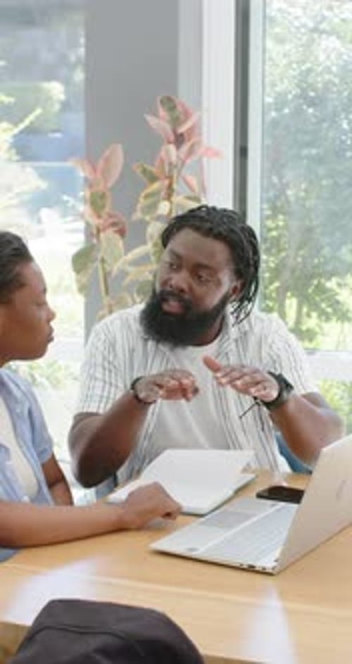 Vidéo verticale : Un homme africain donne des cours à une jeune femme à table faisant des gestes devant un ordinateur portable pendant qu'une étudiante écrit