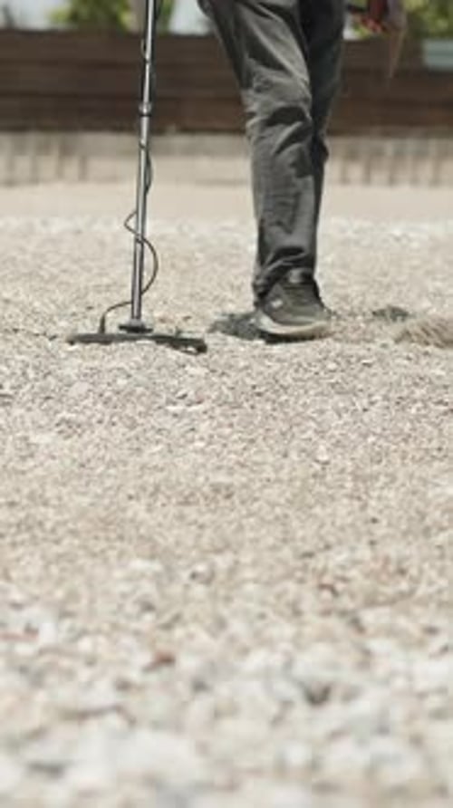 Vertical. A man with a metal detector is searching for lost items on the rocky beach.