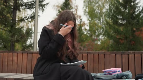 Student Writing Notebook on Bench Outdoor Park Setting with Pine Trees Focused Expression Pen in