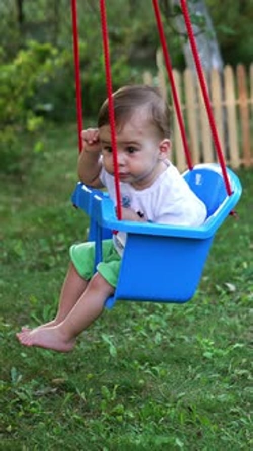Little kid in t-shirt and shorts swaying in a swing. Calm barefoot baby boy in the garden in summer.