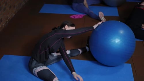 Frauen in Sportbekleidung, die mit weit gespreizten Beinen auf Fitnessmatten sitzen, sich seitlich beugen und Fitbälle bewegen