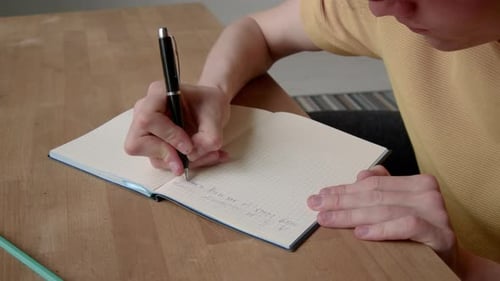 Young Adult Writing in Notebook at Wooden Table
