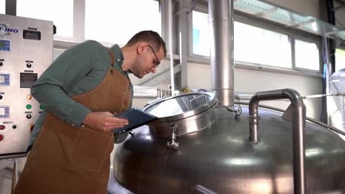 Young brewer working with a modern tablet in a craft beer factory, checking the parameters