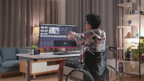 Vista posterior de un hombre en silla de ruedas estirándose mientras editaba y graduaba el color del Vídeo desde un escritorio