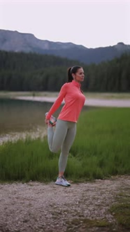 Active female athlete stretching quadriceps muscle near serene mountain lake, showcasing fitness ded