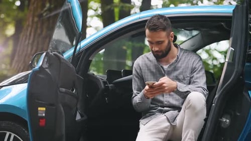 Junger Mann sitzt in dem blauen kleinen Auto und schaut auf das Smartphone. Elektro-Ladestation