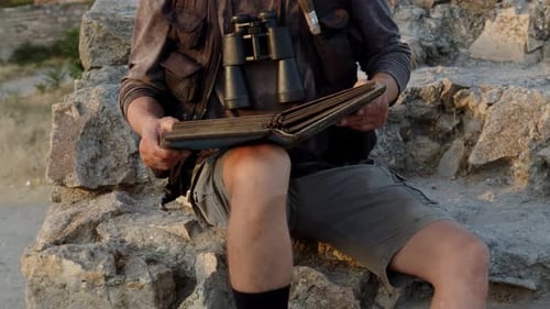 Un homme habillé en chercheur et muni de jumelles est assis sur les rochers et étudie un vieux livre