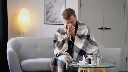 Unhealthy Sad Young Man Sits on the Couch at Home Under a Plaid Sneezing in a Napkin Feels Weak