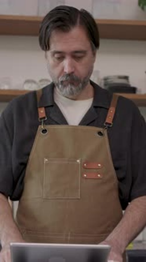 Vídeo vertical de un empleado de café asiático adulto con un delantal marrón usando una tableta digital mientras...