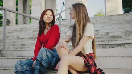 Two Beautiful Girls are Sitting on the Steps in the Park and Talking to Each Other Portrait of Young