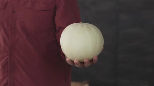 Ein Mann, der mit einer Honigmelone in der linken Hand steht. Er trägt ein magentafarbenes Hemd mit grauem Rücken