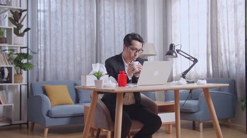 Sick Asian Businessman Drinking Hot Water While Working With Laptop In The Living Room At Home