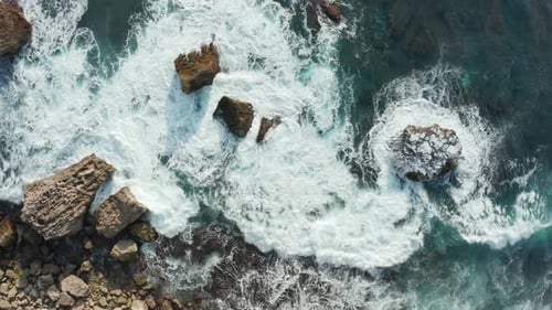Aerial top down view of ocean waves crashing into rocky shore with large rocks. Waves breaking into