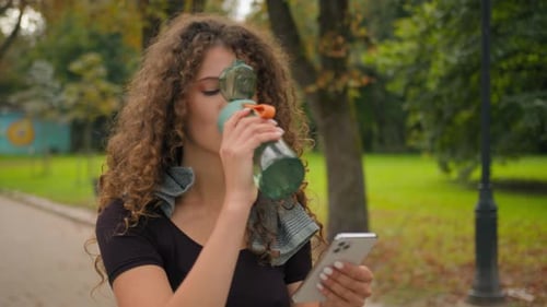 Woman Drinks Water, Uses Phone in the Park