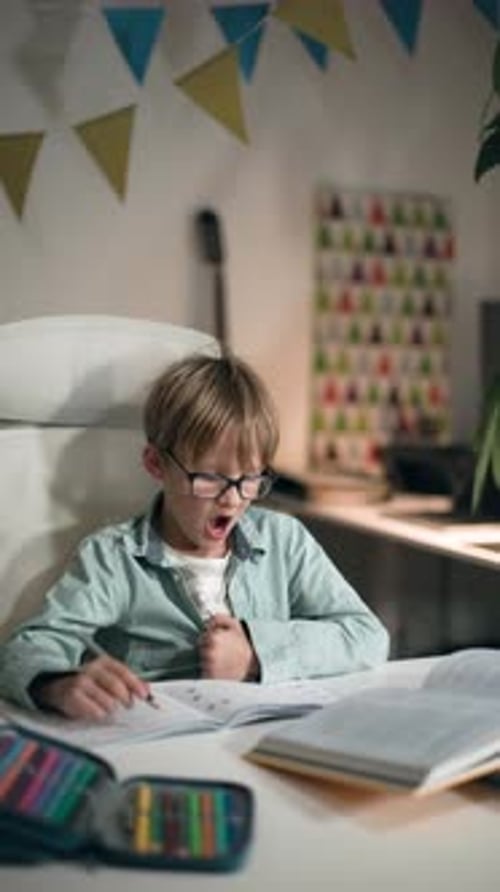 A Child Wearing Glasses Yawns While Sitting at a Table and Doing Homework