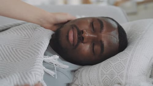 Homme noir malade allongé dans son lit quand il ressent de la fièvre, femme qui s'occupe de lui, gros plan, visage épuisé et