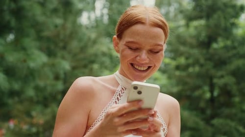 Young Woman Smiles While Using Her Smartphone in a Lush Green Park During Summer