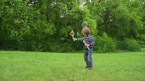 A Happy Boy in a Striped Shirt Creates Large Colorful Bubbles with a Bubble Wand in a Green Park The